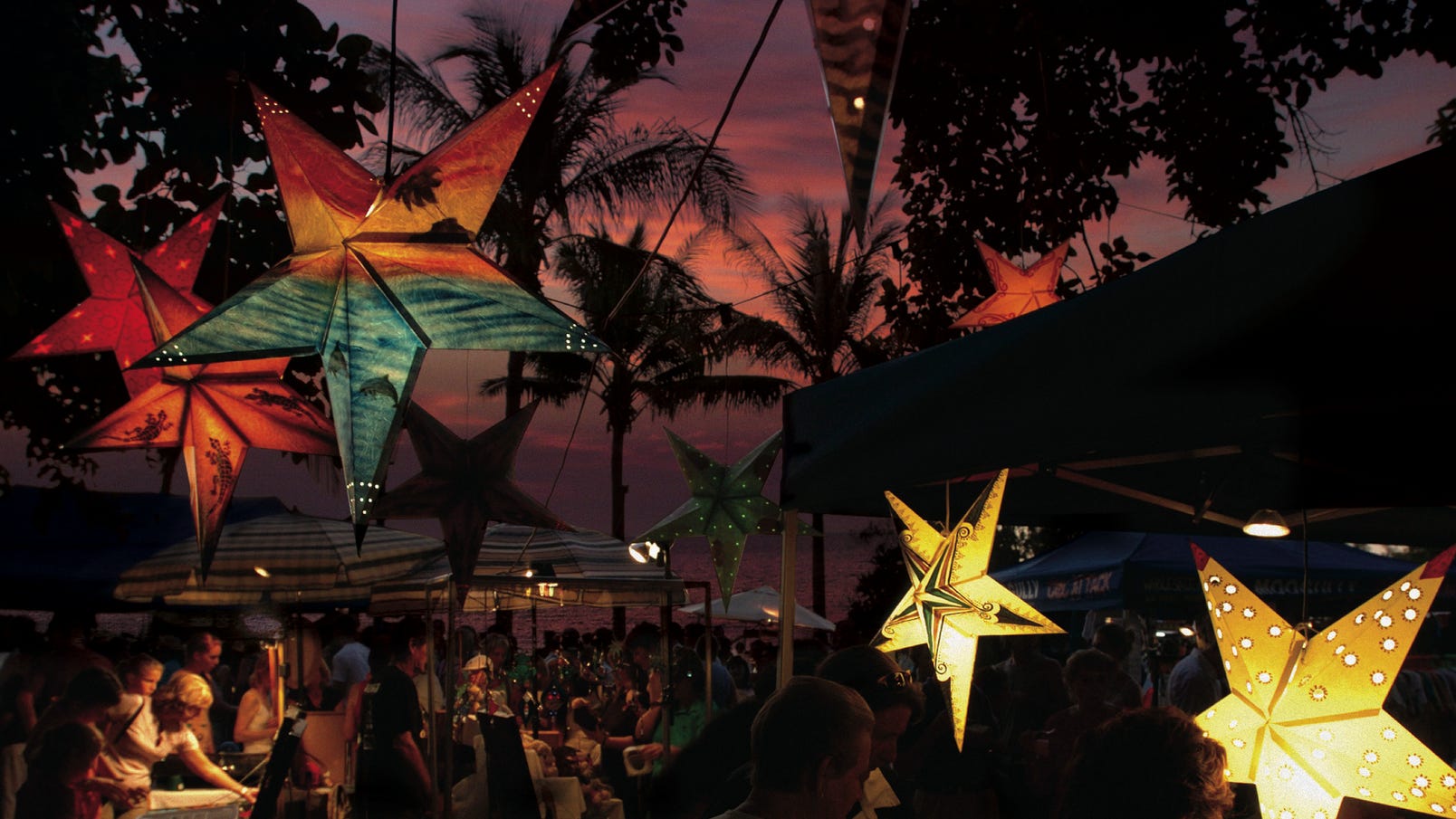 photo of beachside markets at sunset. stalls with people and palm trees dark against the pink and purple sky
